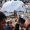 Nell'immagine si vede una tenda bianca in un contesto di distruzione, con rovine e macerie intorno. Un ragazzo è seduto davanti alla tenda, mentre in primo piano ci sono persone che si muovono e tengono stoviglie. L'atmosfera sembra di emergenza e difficoltà.