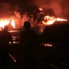 The image depicts two silhouetted figures walking towards a large fire at night, with bright flames and thick smoke rising in the background. The scene conveys a sense of danger and urgency.