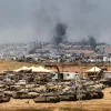 تصویر نشان‌دهنده یک منطقه جنگی با تانک‌ها و دود سیاه از انفجارها در دوردست است.