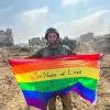 Un soldat souriant tient un drapeau arc-en-ciel sur un terrain dévasté.