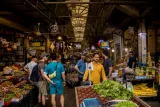 Marché animé, étals colorés de fruits et légumes, gens discutant sous une toiture en métal.