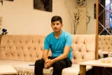 Un jeune homme en t-shirt turquoise est assis sur un canapé beige dans un café.