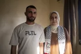 L'image montre deux personnes debout côte à côte dans un intérieur bien éclairé. L'homme porte un t-shirt gris clair avec un logo. La femme, quant à elle, est vêtue d'une robe marron et porte un foulard clair, ainsi qu'une écharpe ornée de motifs traditionnels. Ils semblent se tenir dans une pièce avec des murs clairs et une lumière douce qui entre par une fenêtre.