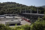 L'image montre une station de péage ou un point de contrôle routier. On aperçoit plusieurs voies de circulation où des véhicules, y compris des camions, attendent. En arrière-plan, on peut voir une structure couverte, probablement destinée à la collecte des péages, ainsi que des pylônes électriques. La scène est entourée de verdure, avec des collines arborées qui le surplombent, sous un ciel clair et ensoleillé.