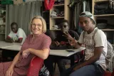 L'image montre un groupe de personnes dans une pièce, probablement une salle de classe ou un espace communautaire. Au premier plan, une femme souriante avec des lunettes et un vêtement à motifs est assise sur une chaise. À l'arrière-plan, deux jeunes hommes sont assis à une table, l'un d'eux regarde son téléphone et l'autre fait un signe de la main. On voit également un troisième homme en arrière-plan. L'ambiance semble conviviale et détendue, avec des vêtements et des accessoires variés dans l'environnement.