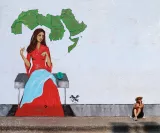 L'image représente une fresque murale captivante. Au centre, une femme vêtue d'une robe rouge est assise, brodant ou tricotant. Elle est entourée d'une carte verte du Moyen-Orient dessinée en arrière-plan. À sa gauche, un globe terrestre est posé. À droite, on aperçoit une personne assise sur le sol, portant un chapeau, qui semble contempler l'œuvre. L'ensemble évoque un mélange d'art et de réflexion sur la culture et la géographie de la région.