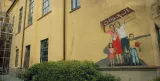 L'image montre un mur extérieur d'un bâtiment peint en jaune, où une fresque colorée est visible. La fresque représente une famille : un homme et une femme, accompagnés de deux enfants. Ils semblent lever les bras en signe de célébration ou de protestation. L'homme porte une boîte sur ses épaules, tandis que la femme est vêtue d'une robe rouge. Les enfants, l'un en chemise et l'autre en robe, semblent joyeux. En arrière-plan, on aperçoit des fenêtres donnant sur l'extérieur. La scène pourrait symboliser des thèmes tels que l'identité, la famille ou le patriotisme.