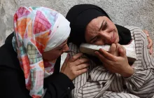 Deux femmes en pleurs, l'une réconfortant l'autre qui tient une chaussure, exprimant une profonde tristesse.