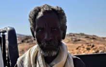 الصورة تظهر رجلاً مسنًا ذا لحية وشعر مجعد، يبدو عليه علامات الزمن والخبرة. عينيه تعكسان نظرة واضحة وقوية، بينما يتواجد في خلفية طبيعة جافة تتضمن صخور وتضاريس غير مستوية. يظهر الرجل بملابس بسيطة، مما يعكس بساطة الحياة في المنطقة التي يعيش فيها. السماء زرقاء والجو يبدو صافياً.
