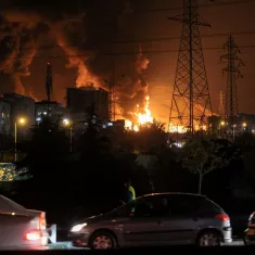En la imagen, hay un gran incendio en una ciudad nocturna, con humo y luces. Autos circulan en primer plano.