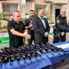 Men in uniform gather around rifles on a table, exchanging greetings and smiles.