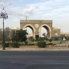 A grand archway with domes, surrounded by greenery and a road, under a cloudy sky.