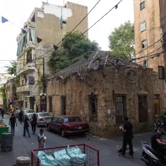 The image depicts a narrow street lined with older buildings, showcasing a blend of urban decay and lively activity. There's a mixture of people, some walking on the street and others gathered in small groups. The buildings have seen better days, with peeling paint and signs of wear. A small, weathered structure with a distinctive roof stands out against the backdrop of larger, more modern buildings. Utility wires crisscross overhead, and there's a hint of greenery from nearby trees. The scene reflects a vibrant yet gritty neighborhood atmosphere.