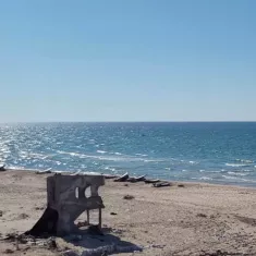 تصویر یک ساحل آرام را نشان می‌دهد. در سمت چپ، بقایای یک ساختار ویران شده قرار دارد. در جلو، شن‌های نرم ساحل و در پس‌زمینه، دریا با امواج ملایم و آبی روشن دیده می‌شود. آسمان صاف و آفتابی است.