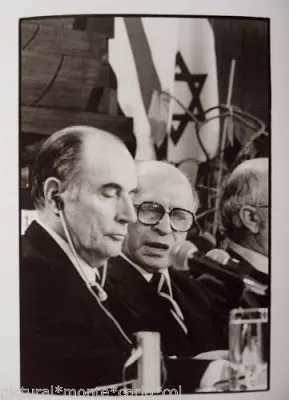 L'image montre deux hommes assis à une table lors d'une conférence. L'un des hommes, portant des lunettes et un costume, semble être en train de parler, tandis que l'autre, avec un regard sérieux, écoute attentivement. En arrière-plan, des drapeaux sont visibles, y compris un drapeau israélien. L'atmosphère semble formelle, ce qui suggère que l'événement a une importance politique ou diplomatique.