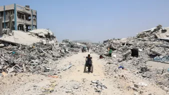 A person in a wheelchair navigates a debris-filled street amid collapsed buildings.