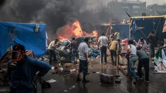 L'image montre une scène de chaos et de destruction. On peut voir des personnes rassemblées autour de flammes, visiblement en train de fuir ou de lutter contre un incendie. La fumée noire s'élève dans le ciel, obscurcissant la vue. Des tentes et des objets sont dispersés dans l'environnement, suggérant une occupation ou une manifestation. La situation semble tendue, avec un sentiment d'urgence parmi les personnes présentes.