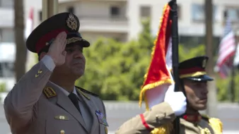 L'image représente deux officiers militaires en uniforme. L'un d'eux, probablement un haut responsable, effectue un salut militaire, tandis que l'autre maintient un drapeau. L'environnement semble être une cérémonie officielle en extérieur, avec des bâtiments en arrière-plan. Les uniformes sont ornés de médailles et de distinctions, reflétant un certain niveau de cérémonie et de formalité.