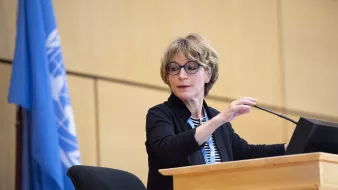 Femme avec des lunettes, parlant devant un podium, drapeau de l'ONU derrière elle.