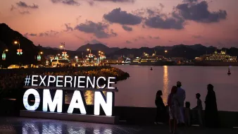 L'image montre une scène au coucher du soleil à Oman, avec des montagnes en arrière-plan et un port où se trouvent des bateaux et des navires de croisière. Au premier plan, il y a un grand panneau lumineux affichant le mot "EXPERIENCE" suivi d'"OMAN". Des personnes se tiennent devant le panneau, admirant la vue sur l'eau. La lumière douce du crépuscule et l'ambiance paisible de la scène créent une atmosphère accueillante.