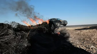 L'image montre un véhicule qui roule rapidement le long d'une route, tandis qu'un feu brûle sur le bord de la route. De la fumée s'élève du feu, qui semble être alimenté par des matériaux tels que des câbles ou des filets. Le ciel est dégagé avec une couleur bleue, et le paysage environnant est aride. On peut voir des débris brûlés au premier plan, ce qui suggère une scène de destruction ou d'accident.