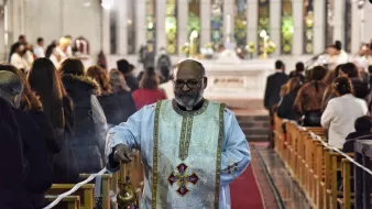 A priest in white robes walks down the aisle of a church during a service.