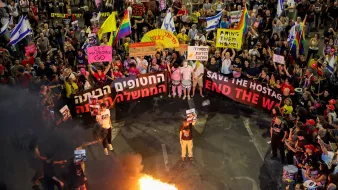 A large protest with colorful banners, people holding signs, and a fire in the center.