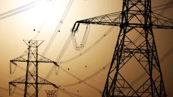 Silhouettes of tall electrical towers against a hazy, orange sunset sky, with power lines connecting them.