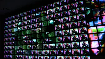L'immagine mostra un ampio muro composto da molteplici schermi televisivi, ognuno dei quali visualizza immagini colorate e astratte. Le immagini si sovrappongono e si mescolano creando un effetto visivo vivace e dinamico. La combinazione di colori e forme sembra generare un'atmosfera futuristica e artistica, dando l'impressione di un'opera d'arte digitale contemporanea.