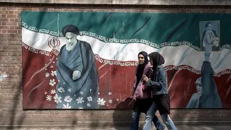 در این تصویر، دو زن در حال قدم زدن در کنار دیواری نقاشی شده هستند که تصویر رهبرایران، امام خمینی، به همراه پرچم ایران و برخی نقوش دیگر بر روی آن نمایان است. زنان به لباس‌های سنتی و مدرنی پوشیده‌اند و در حالی که در حال گفتگو هستند، به سمت دوربین حرکت می‌کنند. دیوار با رنگ‌های سبز، سفید و قرمز پرچم ایران تزئین شده و فضای عمومی شهر را به نمایش می‌گذارد.