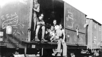 L'image montre un groupe de jeunes hommes assis et debout dans un wagon de train. Ils semblent se trouver dans un environnement rural ou industriel, avec des rails visibles au premier plan. Les murs du wagon sont couverts de graffitis, et l'ambiance générale paraît nostalgique. Les expressions des jeunes varient, allant de la réflexion à l'insouciance. L'image est en noir et blanc, ce qui lui donne un aspect vintage.