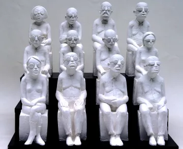 L'image représente une sculpture où plusieurs figures humaines, toutes de couleur blanche, sont assises sur des bancs. Les personnages sont stylisés avec des traits simples et géométriques, portant des lunettes, et semblent avoir des expressions neutres ou pensive. Ils sont agencés en un groupe de dix, disposés en plusieurs rangées, créant une impression d'unité et de contemplation. Les sculptures semblent évoquer des thèmes tels que la solitude, la réflexion ou la condition humaine.