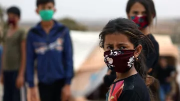 L'image montre un groupe d'enfants, dont une fille au premier plan, portant des masques. La jeune fille a les cheveux bruns et des yeux expressifs, semblant préoccupée. En arrière-plan, d'autres enfants se tiennent, également masqués, dans un environnement qui semble être un camp. L'atmosphère semble chargée d'émotion et de défi. Les couleurs des masques varient, ajoutant une touche de diversité visuelle à la scène.