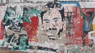 L'immagine mostra un murale su un muro. A sinistra, si può vedere un disegno di una creatura con ali e tratti animaleschi, insieme a elementi astratti e colori vivaci. Al centro, c'è un volto umano stilizzato, con dettagli intricati e un'espressione intensa. Sullo sfondo, si nota un altro personaggio con un gesto di sollevamento delle braccia. La composizione è caratterizzata da una ricca varietà di colori e forme, creando un effetto dinamico e coinvolgente.