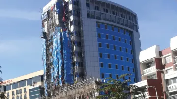 L'image montre un bâtiment en construction. Il s'agit d'une structure de plusieurs étages, avec une façade partiellement terminée, affichant des panneaux de verre de couleur bleue et d'autres en blanc. Le bâtiment est entouré d'échafaudages en métal, et des bâches bleues sont visibles sur certains côtés. À proximité, on aperçoit d'autres bâtiments, probablement de bureaux ou commerciaux, et un ciel bleu en toile de fond. La végétation est également présente au premier plan, ajoutant une touche de verdure à l'environnement urbain.