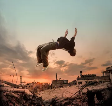 L'immagine mostra un giovane che sembra fluttuare o saltare sopra un paesaggio urbano distrutto, con scarti di costruzione e rovine. Sullo sfondo, si possono vedere nuvole e un cielo al tramonto, che creano un'atmosfera drammatica. La silhouette del ragazzo è evidenziata dalla luce, evidenziando un senso di libertà e movimento in un contesto desolato.