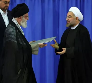L'image montre deux hommes en tenue traditionnelle, se tenant debout l'un en face de l'autre. L'un d'eux porte un turban noir et une cape sombre, tandis que l'autre est vêtu d'une robe noire avec un col ouvert. Ils semblent être en train d'échanger un document ou un dossier, avec des expressions de concentration sur leurs visages. L'arrière-plan est de couleur bleue, ce qui pourrait suggérer un cadre officiel ou un événement marquant.