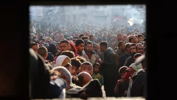 L'image montre une foule dense de personnes, probablement rassemblées dans un espace urbain. Les visages sont variés, et l'atmosphère semble chargée d'émotion et de tension. On peut voir des hommes, des femmes et des enfants, témoignant d'une situation de forte affluence, peut-être liée à un événement particulier comme une distribution d'aide ou une manifestation. Les lumières et l'arrière-plan laissent suggérer un contexte urbain avec des bâtiments visibles. L'image est encadrée par une ouverture, ce qui donne l'impression d'un regard extérieur sur ce rassemblement.