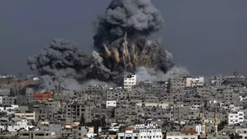 The image depicts a scene of destruction with a large explosion occurring in an urban area. Thick, dark gray smoke billows into the sky, rising above the buildings in the foreground. The skyline is dominated by closely packed structures, showing signs of damage. The overall atmosphere conveys a sense of chaos and devastation, characteristic of an area affected by conflict or military action.