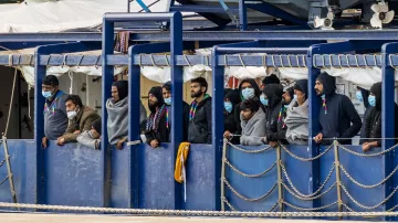 L'image montre un groupe de personnes se tenant sur un bateau. Elles portent des masques et des vêtements semblant chauds, comme des sweats à capuche. Les gens regardent vers l'extérieur, probablement vers la terre ou vers un port. L'environnement semble portuaire, avec des équipements et d'autres structures en arrière-plan. L'atmosphère peut sembler préoccupante ou attendue, reflétant une situation particulière.