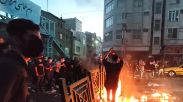 در این تصویر، گروهی از افراد در خیابانی در حال تظاهرات هستند. در مرکز عکس، فردی در حال آتش زدن شیئی است که آتش به‌طور واضحی شعله‌ور شده است. دور تا دور او زباله‌ها و آشغال‌ها پراکنده شده و برخی از معترضان در حال تماشا هستند. بیشتر افراد ماسک به‌دهن دارند و فضای عمومی به نظر شلوغ و تنش‌زا می‌رسد. ساختمان‌های اطراف و نور پایین آسمان نیز به حالت دراماتیک تصویر افزوده است.