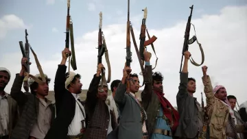 The image shows a group of men holding up rifles in the air. They are standing in a line, with some raising their right arms in a fist while others are focusing on the guns. The setting appears to be outdoors, with a cloudy sky in the background. The men are wearing traditional attire, which varies among them, and there is an overall sense of solidarity and determination in their postures.