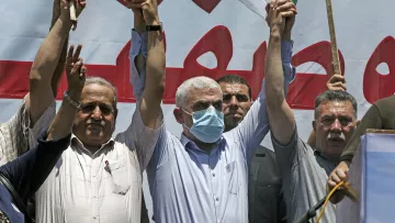 En la imagen se puede ver a un grupo de hombres en una manifestación. Ellos levantan las manos, mostrando un gesto de unidad o celebración. Uno de los hombres en el centro lleva una máscara facial, lo que sugiere que la situación se desarrolla en un contexto donde se requieren medidas de salud. Detrás de ellos hay una pancarta o cartel grande, con texto en árabe y un fondo que parece estar relacionado con la causa por la que están protestando. La atmósfera parece ser de fervor y apoyo colectivo.