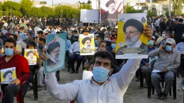 در تصویر، جمعیتی از افراد در یک فضای باز دیده می‌شود که روی صندلی نشسته‌اند و تعدادی از آن‌ها در حال نگه‌داشتن پوسترهایی از یک شخصیت سیاسی هستند. مردم ماسک به‌صورت دارند و در پس‌زمینه، تعدادی پرچم و تابلوهای نمایشی نیز وجود دارد. این جمعیت به نظر می‌رسد که در یک رویداد یا حمایت سیاسی شرکت کرده‌اند.
