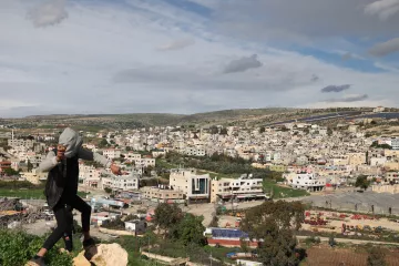در این تصویر، فردی در حال بالا رفتن یا حرکت در مکانی کوهستانی دیده می‌شود. او لباس‌های غیررسمی به تن دارد و در پس‌زمینه، یک شهر بزرگ با ساختمان‌های مختلف و معماری متنوع دیده می‌شود. آسمان ابری است و مناظر طبیعی اطراف، مانند تپه‌ها و درختان، نیز در تصویر قابل مشاهده‌اند. این صحنه ترکیبی از زندگی شهری و طبیعت را نشان می‌دهد.