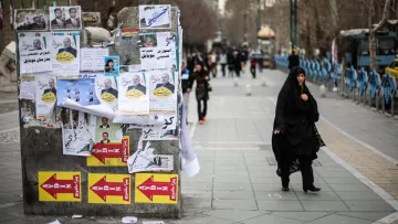 تظهر الصورة مشهداً في منطقة حضرية، حيث تُظهر مجموعة من الملصقات الإعلانية المعلقة على جدار، تتضمن صوراً وعبارات مكتوبة. في المقدمة، توجد امرأة ترتدي حجاباً أسود، تسير في اتجاه معين. الطريق مزدحم بعض الشيء، مع وجود أشخاص آخرين في الخلفية ووسائل النقل. يبدو أن الجو يميل إلى البرودة، ويمكن رؤية الأشجار والبنية التحتية الحضرية.