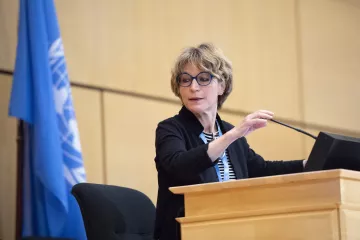 Femme avec des lunettes, parlant devant un podium, drapeau de l'ONU derrière elle.