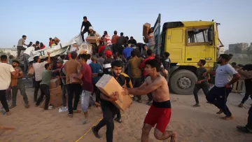 در این تصویر، گروهی از افراد در حال جابجایی و بارگیری جعبه‌ها از یک کامیون بزرگ هستند. برخی از افراد با جعبه‌ها به سمت کامیون می‌روند و دیگران در حال خارج کردن جعبه‌ها از کامیون به زمین هستند. به نظر می‌رسد این فعالیت در فضای باز و در یک منطقه شنی در حال انجام است. افراد در حال همکاری و تلاش برای حمل و نقل بسته‌ها هستند و جو تصویر به شدت忙 و پر از فعالیت به نظر می‌رسد.
