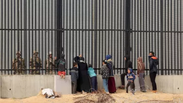The image depicts a group of people, including women and children, standing in front of a tall, barred fence. On the other side of the fence are soldiers or guards, who appear to be watching the group. The scene conveys a sense of separation and tension, with barbed wire in the foreground emphasizing the barriers between the two sides. The individuals on the left seem to be engaging with the guards, possibly seeking assistance or communication. The overall atmosphere suggests a complex and challenging situation, likely related to migration or security issues.