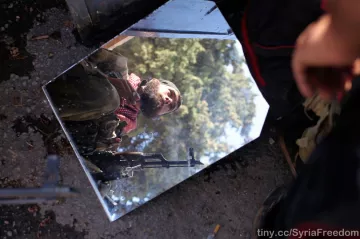 L'image montre un miroir fragmenté reposant sur le sol, reflétant l'image d'un homme en uniforme avec un fusil. À l'arrière-plan, on voit des arbres, suggérant un environnement naturel ou une zone de conflit. Le reflet donne une perspective intrigante sur la scène. L'homme semble vigilant, peut-être en attente ou en préparation pour une opération.